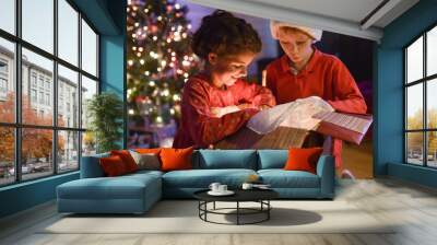 2 children opening a gift in front of the christmas tree lit up, Wall mural