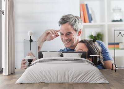 Cheerful couple sitting on a white couch at home using a tablet Wall mural