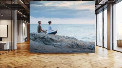 Couple practicing yoga at sunrise on the beach facing the sea Wall mural