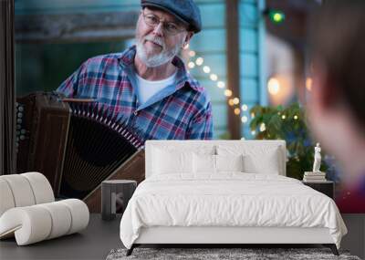 In the evening, a bearded musician with a cap plays accordion in front of a bar outside in front of the spectators. Wall mural