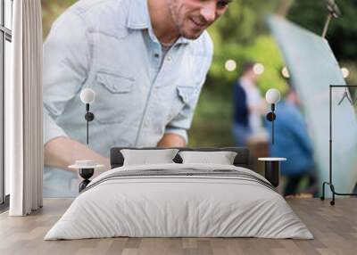 in the summer,  a handsome man in his forties prepares a barbecue for his friends gathered around a table in the garden Wall mural