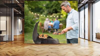 in the summer,  a handsome man in his forties prepares a barbecue for his friends gathered around a table in the garden Wall mural