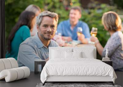 Looking at the camera, a man in his forties sits at a table in the garden with his friends for dinner and have good time Wall mural