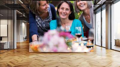 Summertime. Group of friends gathered around a table in the garden. Three beautiful women in their 40s pose for the camera. Wall mural