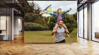 Father carrying son playing with kite in park Wall mural