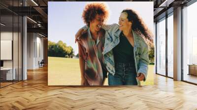 Two young girlfriends bonding outdoors, laughing and embracing on a sunny day in a green park landscape Wall mural