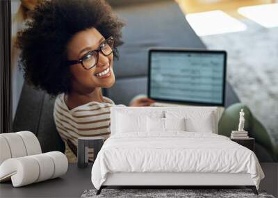 No better way to work than to work from home. Portrait of a cheerful young woman working on her laptop while being seated comfortably on a couch at home. Wall mural