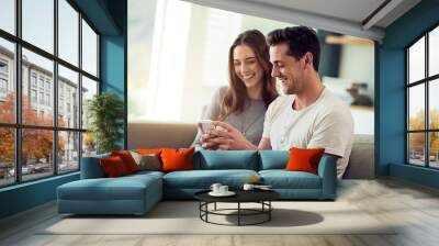 Sharing is caring. Shot of a happy young couple using a phone together on the sofa at home. Wall mural