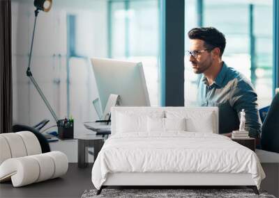 The successful stay consistent. Shot of a focused young businessman using a computer at his desk in a modern office. Wall mural