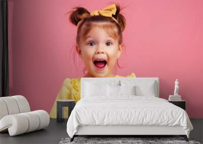 Excited little girl in a yellow dress with a matching bow on a pink background. Happy child with an open mouth expression. Childhood, joy, playful, portrait concept Wall mural