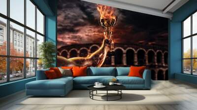 Muscular man arm holding burning trophy cup, antique colosseum on background. Wall mural