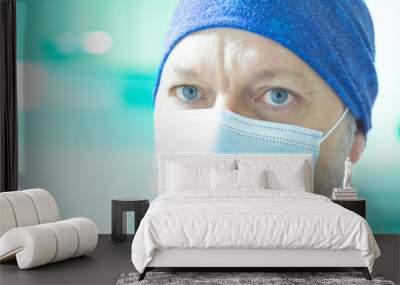 Close-up of a doctor with blue eyes and a surgical mask looking straight into the camera in a primary care medical consultation room of a health center with a green background out of focus Wall mural