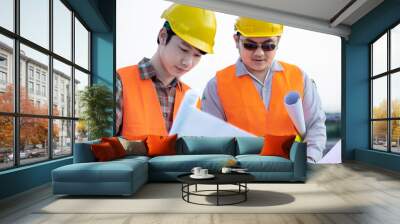 Two Asian engineers or technicians wear a yellow helmet with an orange reflective jacket and stand to hold a blueprint with a smile. Both consult the rooftop or construction site. Wall mural