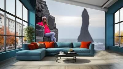 Beautiful girl in pink jacket sits on basalt columns with big rock (troll fingers) on famous Reynisfjara black beach, Iceland Wall mural