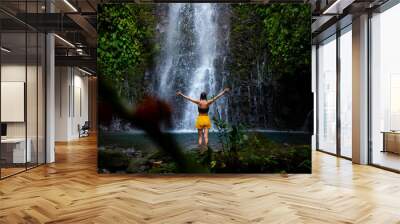 brave girl stands in front of a mighty waterfall with her hands raised in the air; celebration of a successful climb over a waterfall; tropical waterfall in Costa Rica; hidden gems of costa rica Wall mural