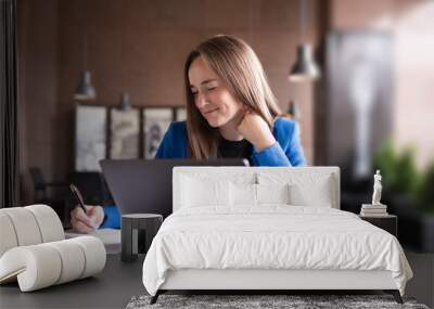 Beautiful girl with a laptop smiles and writes in a notebook. Wall mural