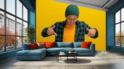 An excited young Asian man, dressed in a beanie hat and casual shirt, points his finger downward toward an empty copy space while standing against a yellow background Wall mural