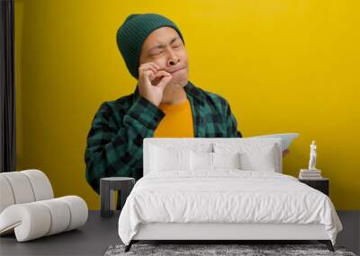 Asian man signals a delicious gesture with an OK sign while holding an empty plate, expressing satisfaction and happiness, possibly due to having enjoyed delicious food, isolated on yellow background Wall mural