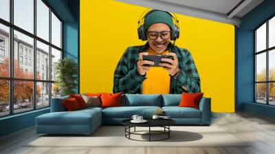 Excited young Asian man, wearing headphones, a beanie hat and casual shirt, engages in playing an online mobile game on his phone while simultaneously live broadcasting the gameplay on the internet Wall mural