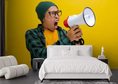 Young Asian man in a beanie hat and casual shirt shouts through a megaphone, expressing annoyance and frustration with angry yelling. Isolated on a yellow background Wall mural