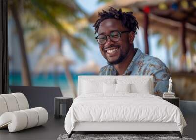 A happy black man in his thirties working on a laptop at a tropical beach bar, using the computer for remote work while vacationing at an exotic island resort with palm trees and blue ocean water. Wall mural