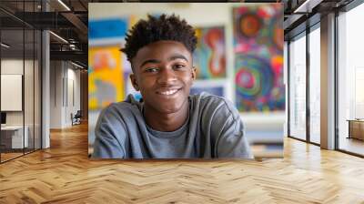 African American teenage boy sitting at his desk in a middle school classroom, smiling and looking directly into the camera. The room is filled with colorful decorations on the wall behind him. Wall mural