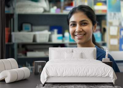 Attractive young Indian nurse wearing blue scrubs, smiling and standing in front of medical equipment on shelves behind her with colors and stationery in a bright light office space. Wall mural