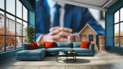 Businessman sitting at a desk with a miniature house and stacks of coins, representing real estate investment and financial planning. Wall mural