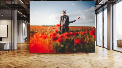 Beautiful young blonde girl dreeming and holding a poppies bouquet in poppy field at sunset in leather jacket from behind Wall mural