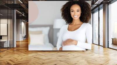 A pregnant woman smiling while holding her belly, sitting in a bright, cozy bedroom. Wall mural