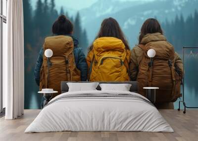 Three female hikers with bright yellow backpacks walking through a green meadow towards misty mountains, enjoying a peaceful hike in a scenic landscape. Wall mural