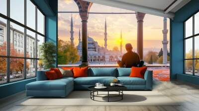 Young man reading the Quran in front of an ancient mosque at sunrise, with blue dome and white spires visible through the window. Wall mural