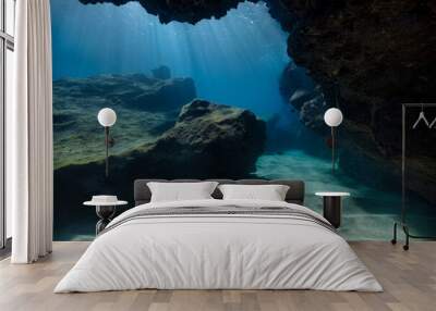 An underwater scenic view from inside a cave as the sun penetrates the water, raining sun rays down on the sandy and rocky sea floor Wall mural