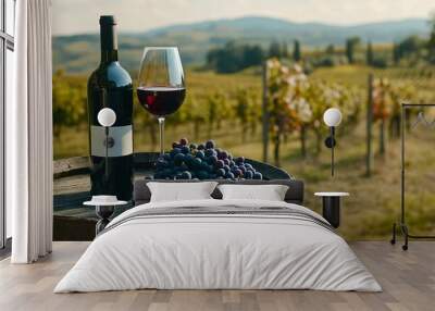 A bottle of red wine and a glass of wine sit on a wooden barrel surrounded by fresh grapes in a picturesque vineyard with rolling hills in the background. Wall mural