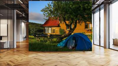 Pretty rural cottage with a blue tent in front of house. Idyllic mountain landscape with bench under the tree. Wall mural