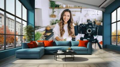 A young woman food blogger cooking salad in front of smartphone camera while recording vlog video and live streaming at home in kitchen. Wall mural