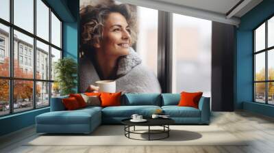 Portrait of happy middle aged woman in cozy sweater holding a cup of hot drink and looking trough the window, enjoying the winter morning at home, side view Wall mural