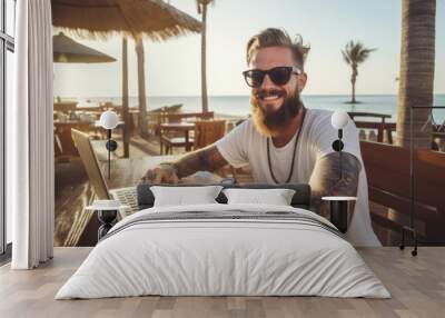 Smiling handsome hipster man with tattoos sitting and working on his laptop in the beach bar Wall mural