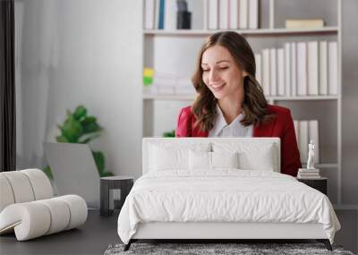 Concept of business working, Businesswoman wearing red suite sitting while writing and checking business analytic document  report  with computer laptop, smartphone on desk in her workstation. Wall mural