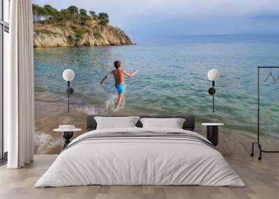 child runs along the shore towards the sea splashing water to take a dip in a beach of crystal clear turquoise waters with a cliff in the background Wall mural