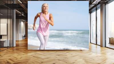 Mature woman running along the shore of the beach. Older female doing sport to keep fit Wall mural