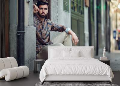 Young bearded man, model of fashion, sitting in an urban step wearing casual clothes. Wall mural