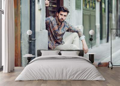 Young bearded man, model of fashion, sitting in an urban step wearing casual clothes. Wall mural