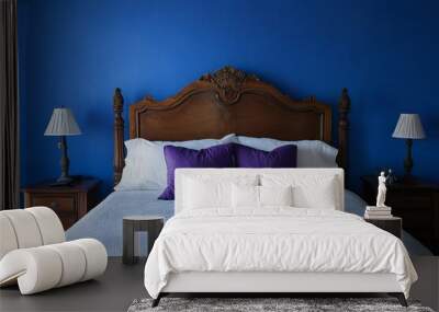 A bedroom with a blue wall and a white bed with a wooden headboard. The bed is made with white sheets and has violet pillows. There are two nightstands on either side of the bed, both with a lamp Wall mural