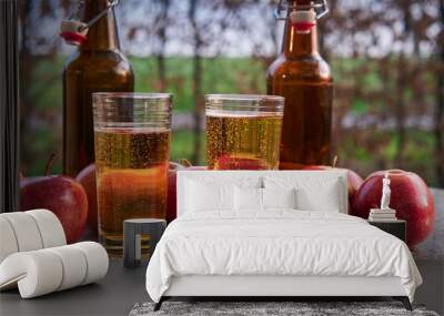 Close up Picture on two glasses or jars full of organic sparkling apple cider and two rustic bottles with red riped apples on stone garnit table in the garden restaurant during summer sunny evening. Wall mural