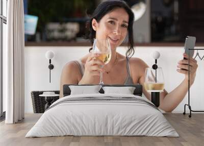 Adult woman sitting in an outdoor restaurant taking a selfie with her phone while holding a glass of wine looking at camera Wall mural