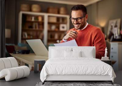 A smiling male remote worker comparing a document in his hand to the one on his laptop. Wall mural