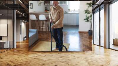 Bearded senior man cleaning carpet with vacuum cleaner at home. Senior man vacuum cleaning home. Wall mural