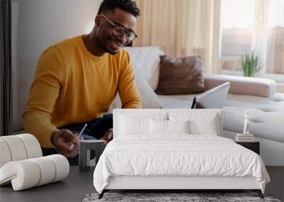 Cropped shot of a handsome young businessman sitting alone in his home office and working on laptop computer at home. Smiling black man using laptop in living room. Businessman using laptop at home. Wall mural