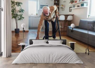 Senior man using his vacuum cleaner. Bearded senior man vacuuming carpet, male cleaning house using vacuum keeping home clean, housekeeper, everyday life concept. Wall mural